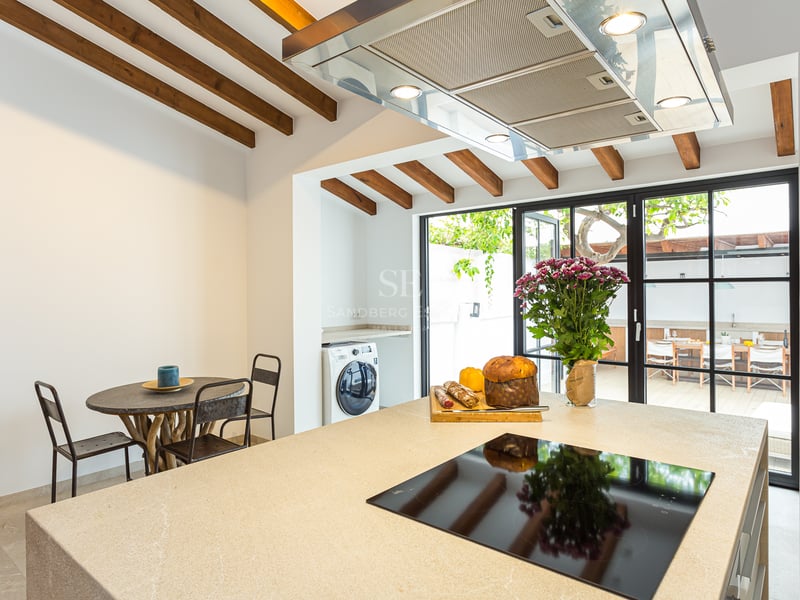 Une cuisine moderne avec un îlot en pierre beige, plaque à induction et poutres rustiques donnant sur une terrasse.