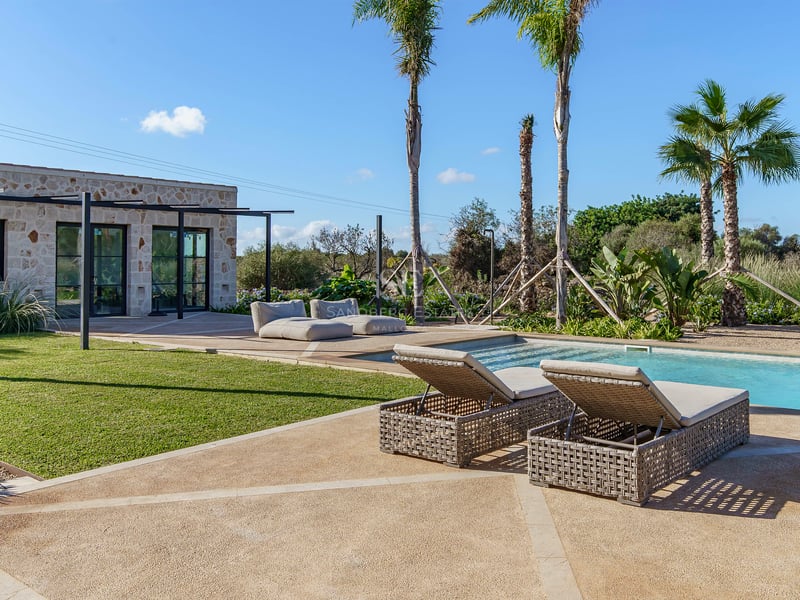 Piscine de luxe avec bains de soleil, palmiers et pavillon en pierre naturelle sous un ciel dégagé.