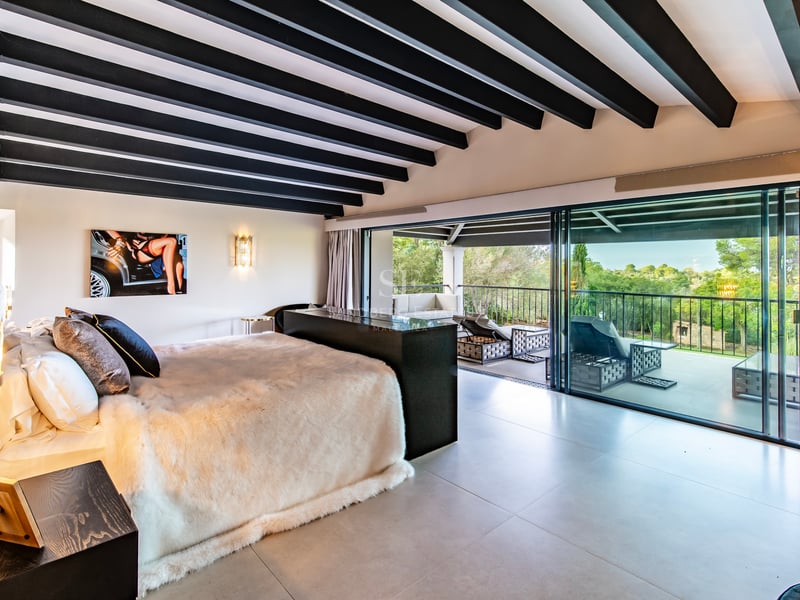 Chambre de maître moderne avec poutres sombres et baies vitrées ouvrant sur une terrasse avec vue sur la nature.