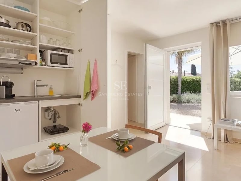 Kitchenette blanche moderne et table à manger avec porte ouverte sur un jardin ensoleillé et un palmier.