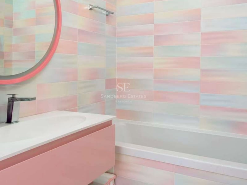 Salle de bains avec baignoire, meuble sous vasque rose et carrelage mural irisé aux couleurs douces.