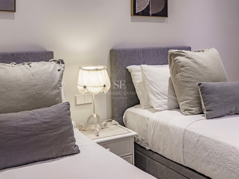 Deux lits jumeaux avec têtes de lit rembourrées grises, draps blancs et une lampe sur une table de chevet entre eux.