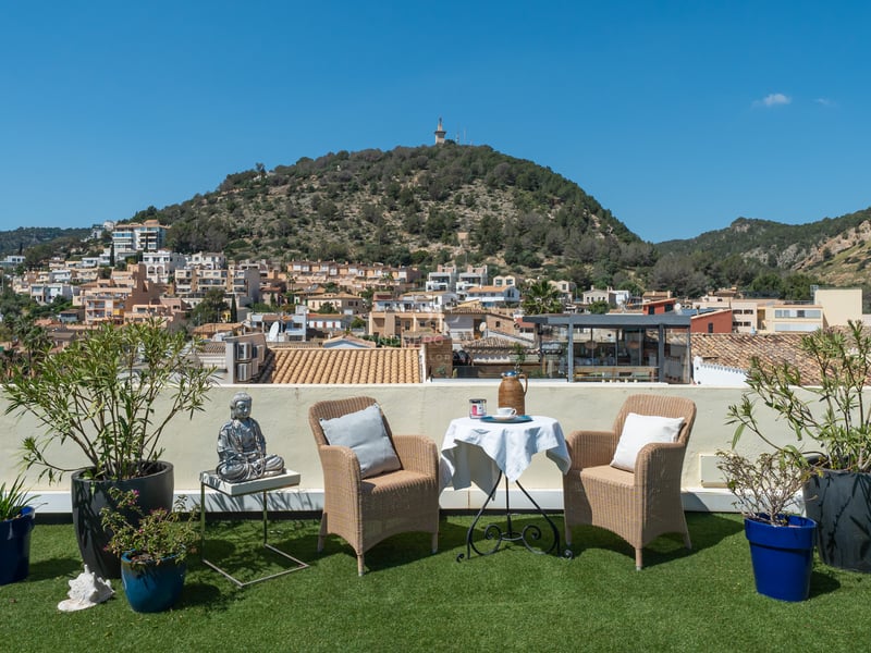 Terrasse sur le toit avec fauteuils en osier, table et plantes donnant sur un village et une colline méditerranéenne.