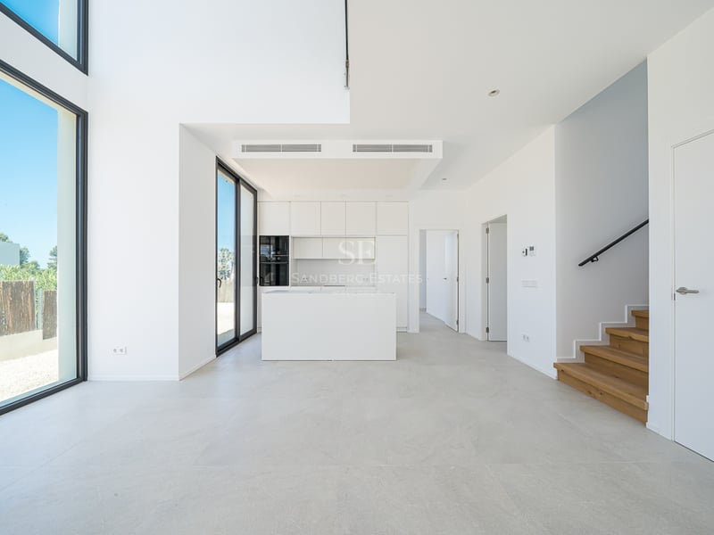 Cuisine blanche minimaliste avec îlot, escalier en bois et grandes baies vitrées du sol au plafond.