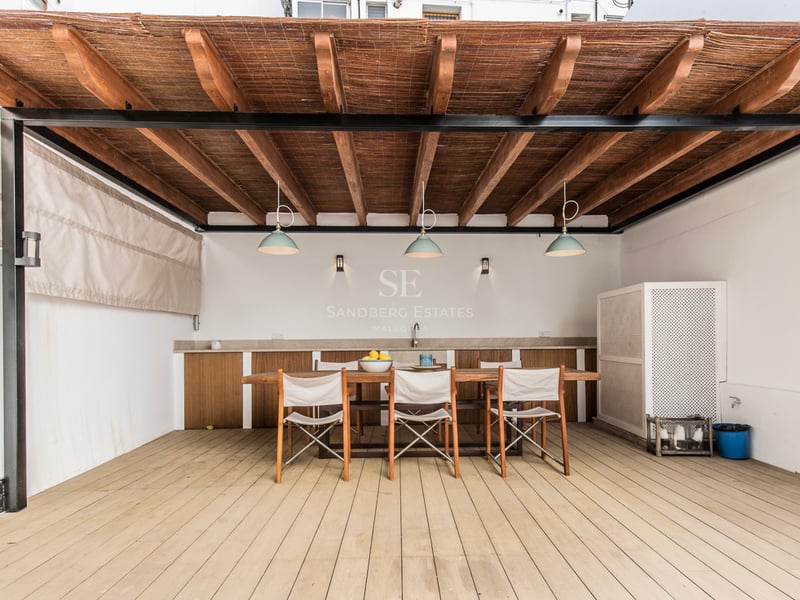 Salle à manger extérieure ombragée avec une grande table en bois, des chaises et une pergola avec kitchenette.