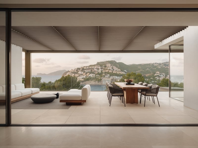 Terrasse couverte moderne avec table en bois, canapés blancs et vue sur mer à travers des garde-corps en verre.