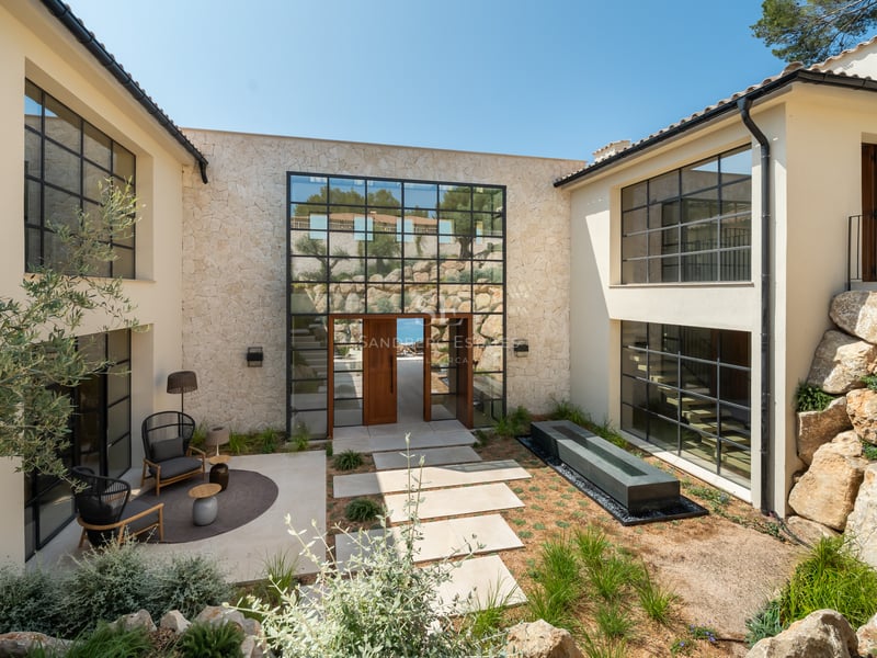 Entrée de villa moderne avec façade en pierre, grandes fenêtres à carreaux et patio paysager avec salon extérieur.