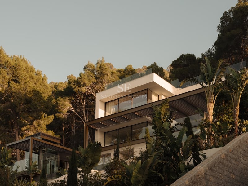 Villa blanche moderne à plusieurs niveaux avec balcons en verre, entourée de pins et de plantes tropicales.