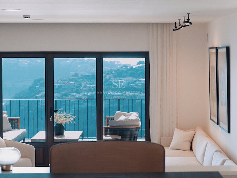 Salon lumineux avec canapé blanc et grandes baies vitrées donnant sur une colline et du mobilier de balcon.
