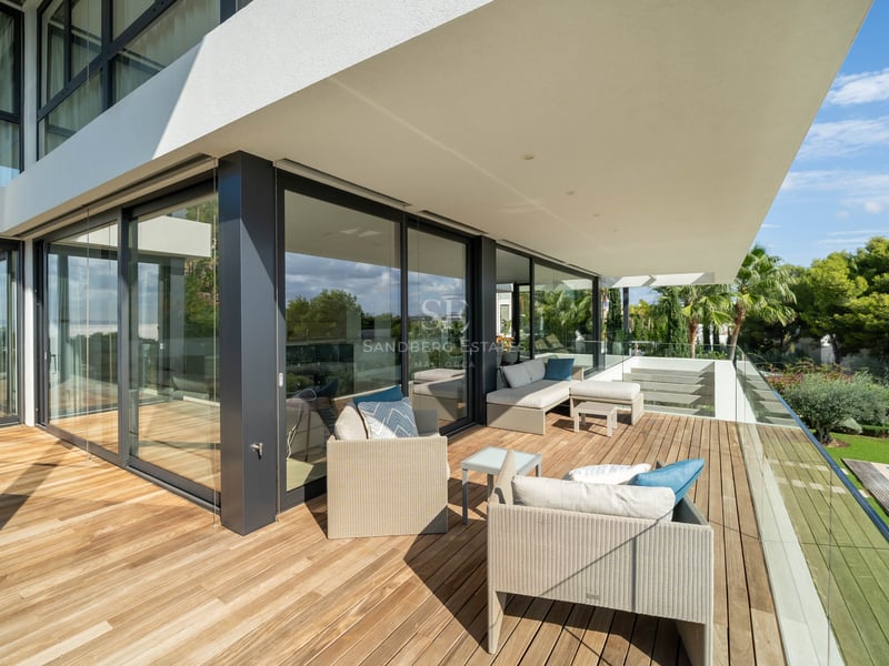 Terrasse spacieuse en bois avec mobilier lounge et garde-corps en verre surplombant une piscine et un jardin.