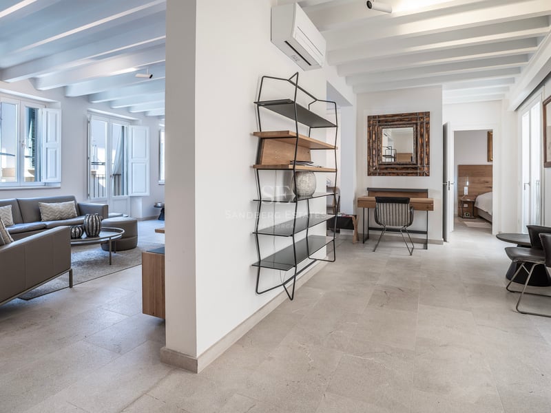 Grand salon moderne avec canapé en cuir gris, sols en pierre, poutres apparentes et étagère minimaliste.
