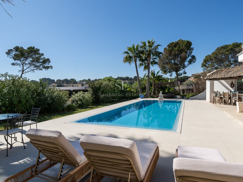 Piscine turquoise entourée de dalles blanches, chaises longues, palmiers et villa méditerranéenne sous un ciel bleu.