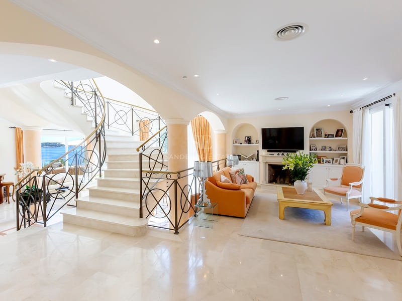 Salon spacieux avec grand escalier en marbre, fenêtres cintrées avec vue mer et cheminée classique.