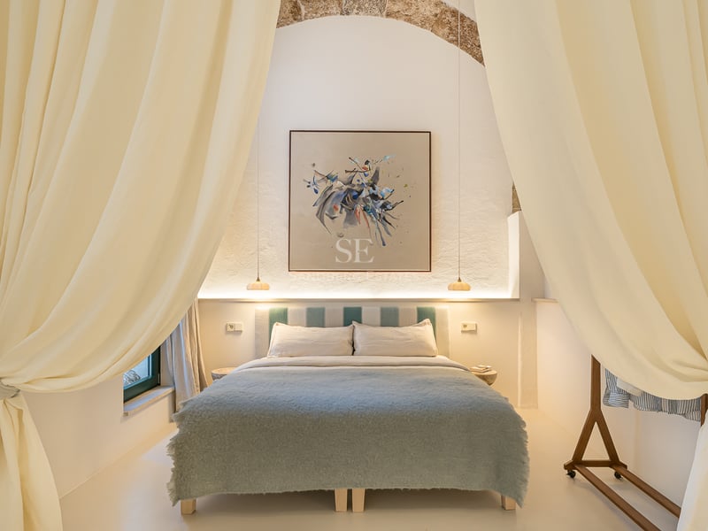 Chambre élégante avec grand lit, rideaux crème, arche en pierre apparente et tableau abstrait moderne.