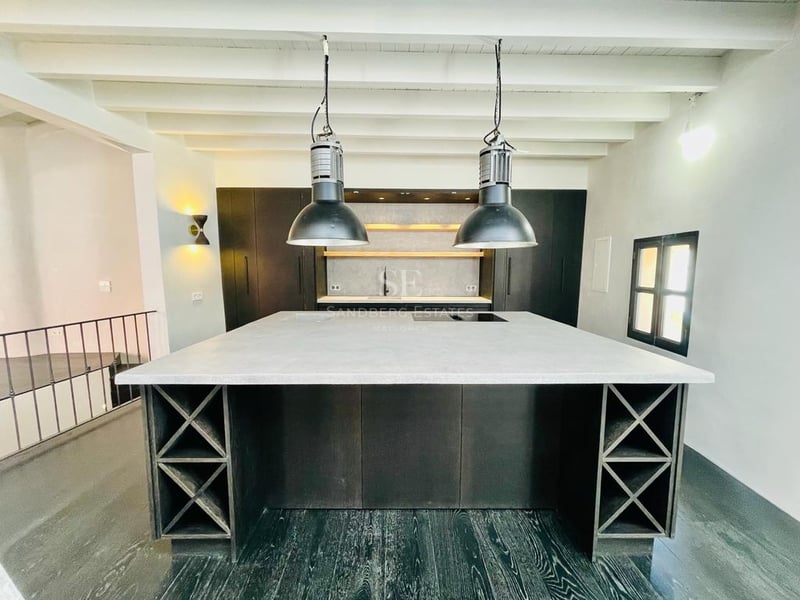 Cuisine moderne avec grand îlot gris, casiers à vin, suspensions industrielles et plafond à poutres blanches.