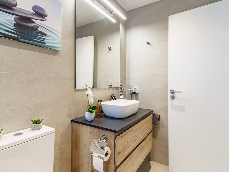 Salle de bain moderne avec vasque à poser, meuble en bois, murs carrelés beige et miroir avec éclairage LED.