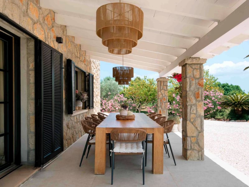 Grande table en bois sous un porche avec colonnes en pierre, luminaires en rotin et vue sur un jardin fleuri.