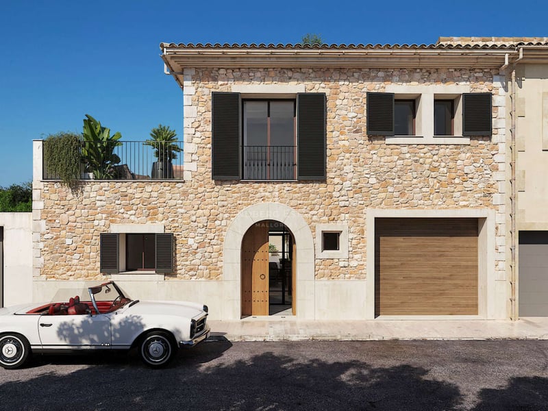 Maison de village en pierre avec porte voûtée, volets sombres et cabriolet Mercedes classique blanc stationné devant.