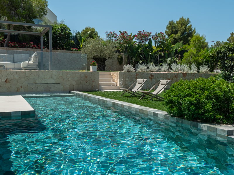 Piscine moderne avec eau turquoise, murs en pierre naturelle et jardin paysager sous un ciel bleu.