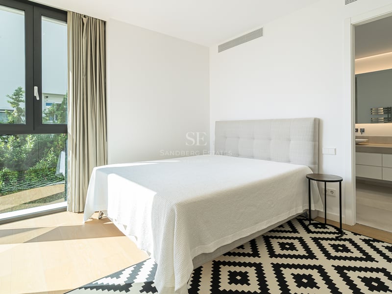 Chambre moderne avec lit blanc, tapis géométrique, grande fenêtre sur jardin et salle de bain attenante.