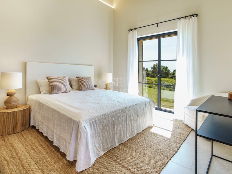 Dormitorio luminoso con cama blanca, alfombra de yute y ventanales con vistas a un entorno verde.