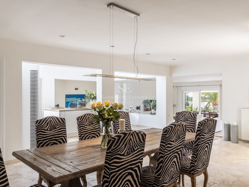 Table à manger en bois rustique, chaises à motif zébré et sols en travertin dans une villa lumineuse.