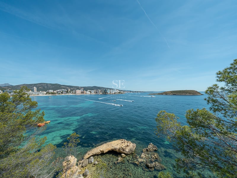 Vue d'une baie aux eaux turquoises avec des bateaux, entourée de pins et d'une ville côtière au loin.