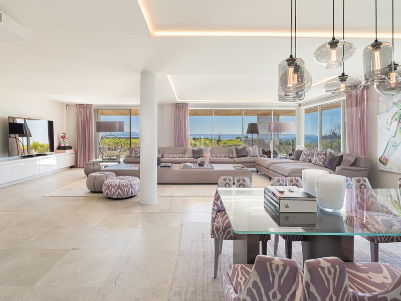 Salon ouvert moderne avec canapé d'angle mauve, table de salle à manger en verre et grandes baies vitrées sur la mer.