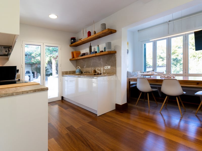 Cuisine blanche moderne avec plans de travail en pierre, étagères en bois et coin repas près de la fenêtre.