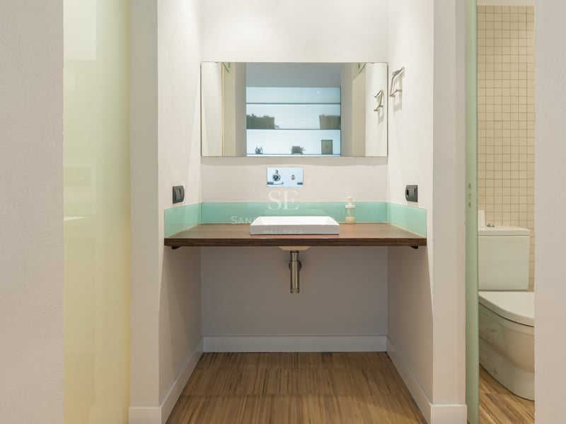 Salle de bain avec plan de travail flottant en bois, vasque carrée blanche, miroir et crédence en verre turquoise.