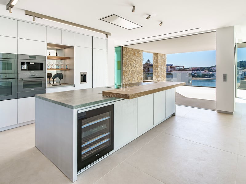 Cuisine minimaliste moderne avec îlot central, cave à vin et baies vitrées ouvrant sur une terrasse vue mer.