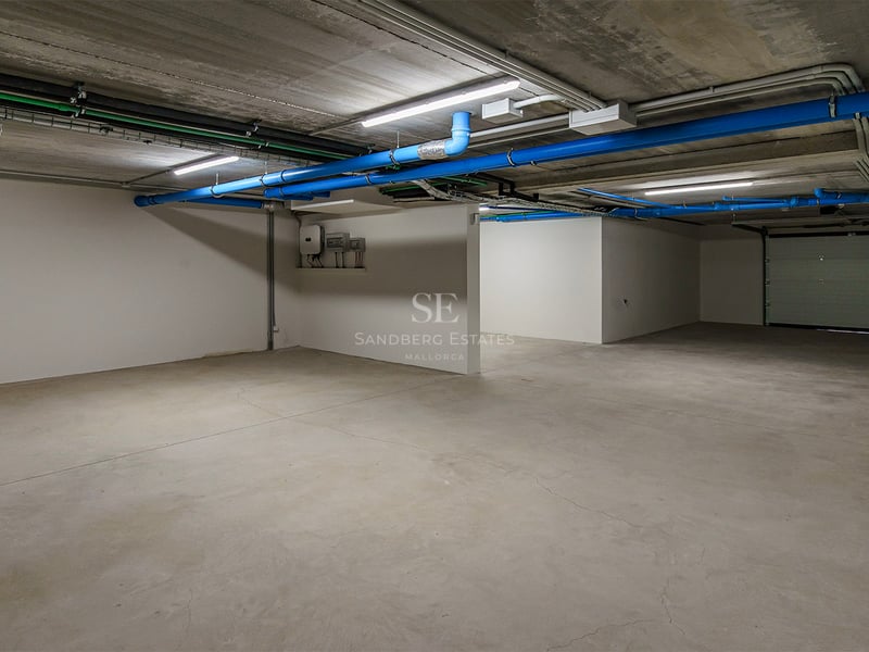 Garage souterrain vide avec sols en béton, tuyaux bleus et murs blancs sous éclairage LED.