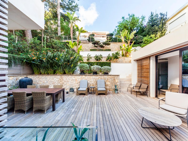 Grande terrasse en bois avec mobilier en osier devant un mur de pierre à plusieurs niveaux et plantes tropicales.