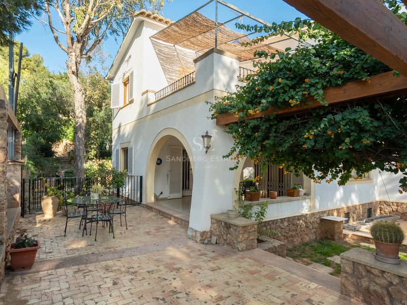 Patio ensoleillé en dalles, pergola en bois fleurie et façade de villa blanche avec arches.