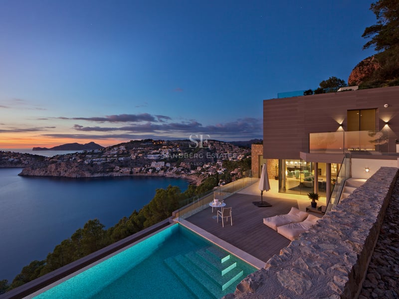 Terrasse d'une villa moderne avec piscine à débordement surplombant une baie au coucher du soleil.