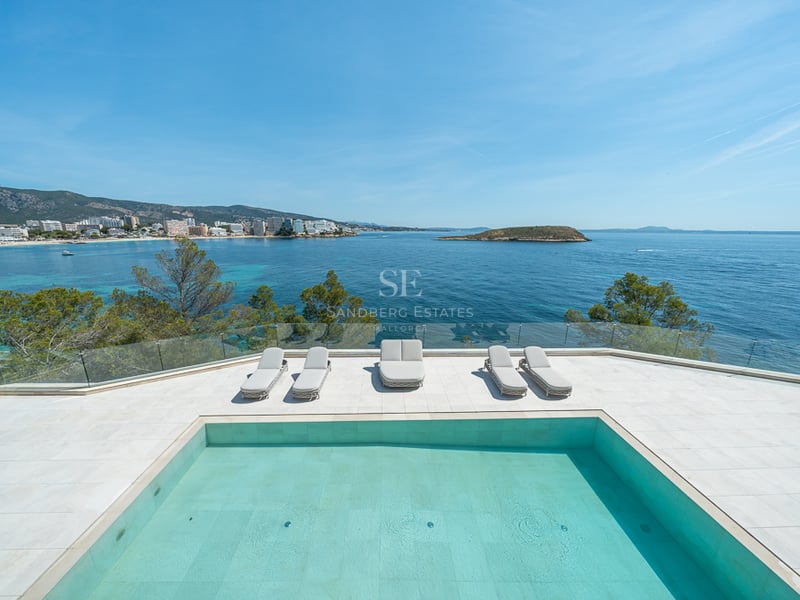 Piscine moderne et chaises longues sur une terrasse blanche surplombant une baie turquoise et le littoral.