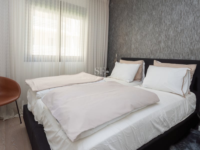 Chambre moderne avec lit sombre, mur texturé gris, fauteuil en cuir tabac et rideaux blancs toute hauteur.