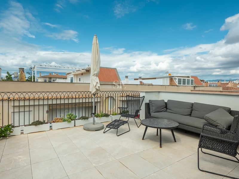 Terrasse de toit avec canapé gris, chaises noires et parasol blanc sous un ciel bleu.