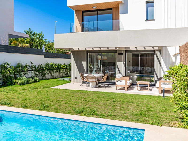 Modern vit villa med turkos pool, grön gräsmatta och möblerad terrass under en klarblå himmel.