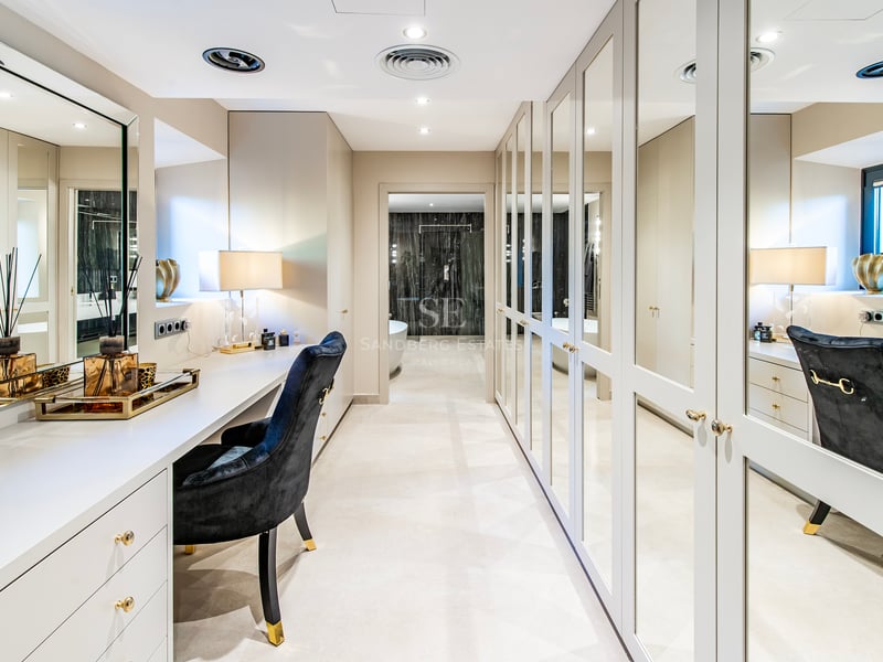 Dressing lumineux avec placards à miroir, une coiffeuse blanche et une chaise en velours noir menant à la salle de bain.