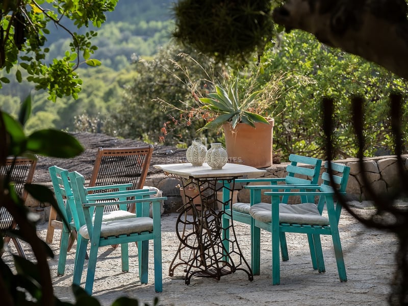 Table en marbre vintage et chaises en bois bleu canard sur un patio en pierre entouré de verdure méditerranéenne.