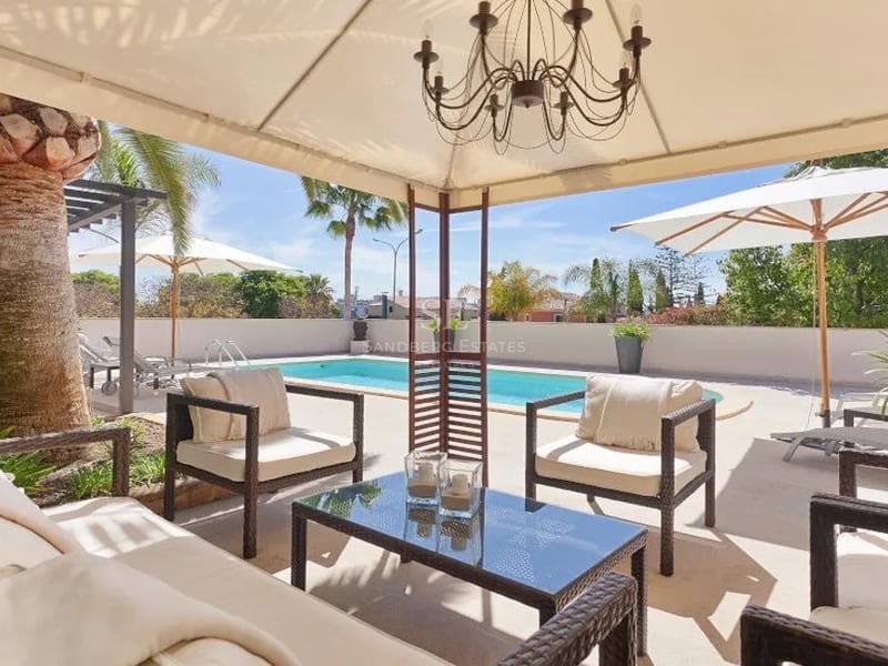 Terrasse ombragée avec mobilier en rotin et lustre, vue sur une piscine privée et des palmiers sous un ciel bleu.