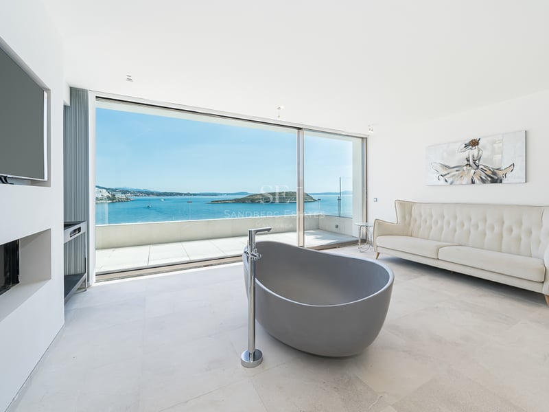 Salle de bain moderne avec baignoire îlot grise, canapé crème et baies vitrées offrant une vue panoramique sur la mer.