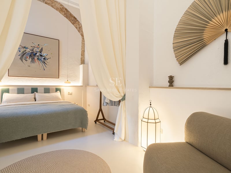 Chambre de luxe avec grand lit, arche en pierre d'origine, rideaux fluides et décor méditerranéen minimaliste.