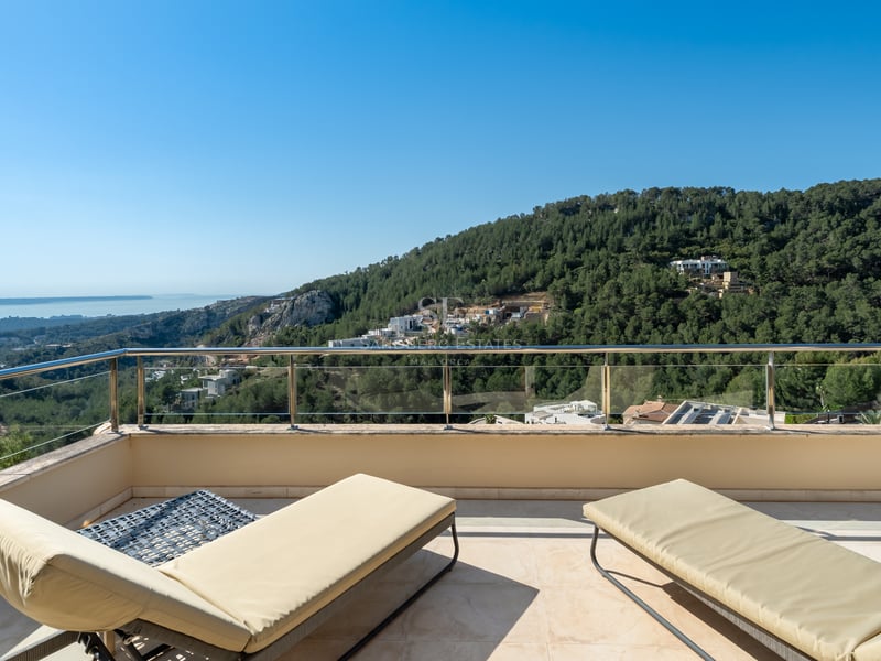 Deux chaises longues crème sur une terrasse en pierre avec garde-corps en verre surplombant une vallée et la mer.