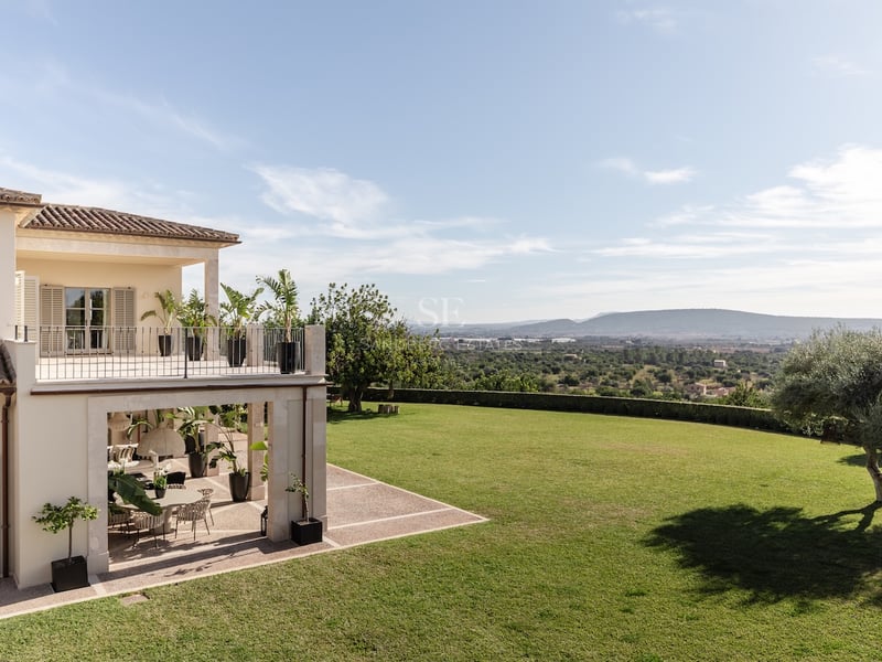 Villa crème avec vaste pelouse, terrasse ornée de palmiers et vue panoramique sur les montagnes par temps clair.