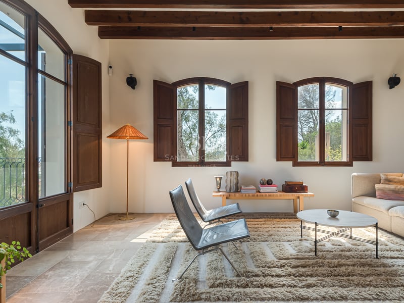 Salon lumineux avec sol en pierre, poutres en bois sombre, fenêtres cintrées avec volets et mobilier moderne.