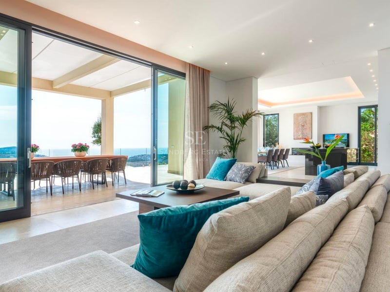 Vaste salon moderne avec canapé beige, baies vitrées toute hauteur et terrasse donnant sur la mer.