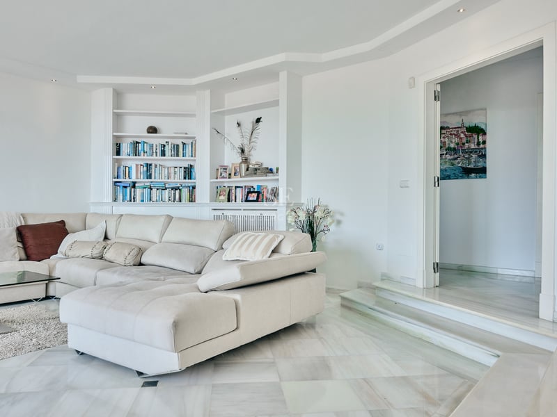 Salon contemporain avec sol en marbre blanc, grand canapé d'angle crème et bibliothèque intégrée blanche.
