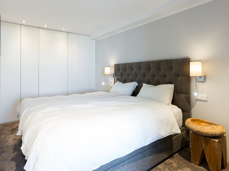 Dormitorio contemporáneo con cama blanca, cabecero gris acolchado, armarios blancos y mesita de madera tallada.